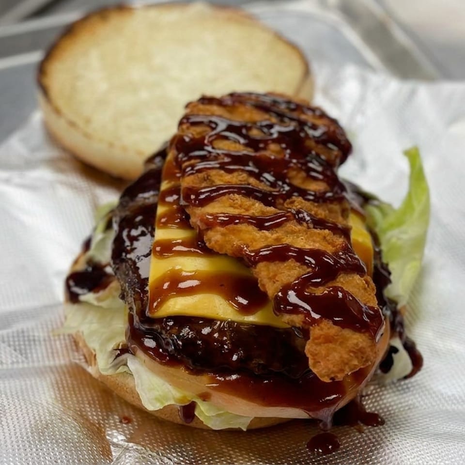 Loaded cheeseburger with lettuce, crispy fried topping, and glossy barbecue sauce on a napkin-lined metal tray.