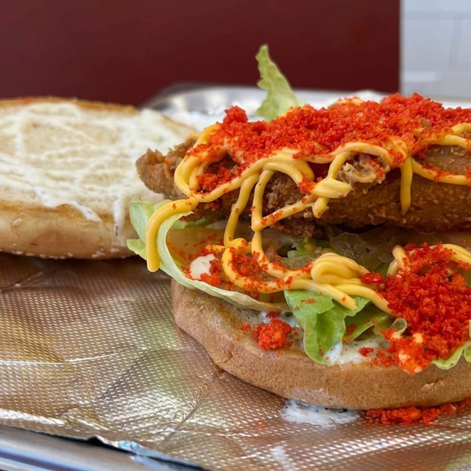 Open sandwich with a crispy meat patty, lettuce, creamy sauce, yellow drizzle, and red crumb topping on foil.