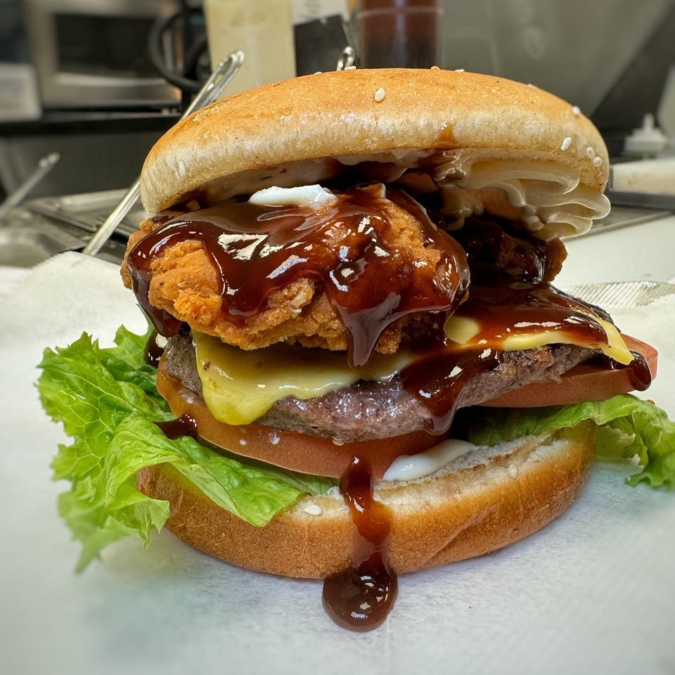 Double cheeseburger topped with crispy fried chicken, lettuce, tomato, creamy sauce, and glossy barbecue sauce on a sesame bun.